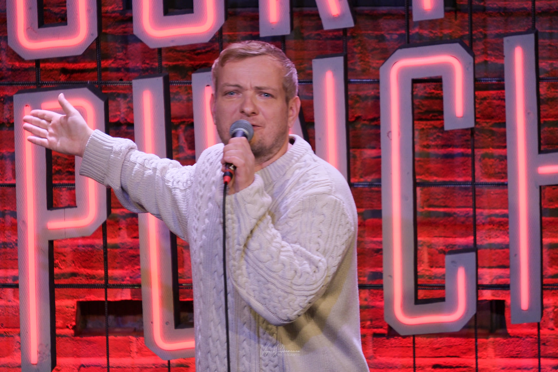 Shane Callaghan performing stand-up, mic in hand, against a red-lit club wall